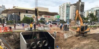 grande peça de sistema pluvial içada por um guindaste gigante ao lado do calçadão da beira-mar e operários em volta