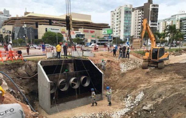 grande peça de sistema pluvial içada por um guindaste gigante ao lado do calçadão da beira-mar e operários em volta