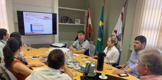 vereadores e outras pessoas reunidas em volta de uma mesa em um escritório olham para a tela de uma tv na ponta