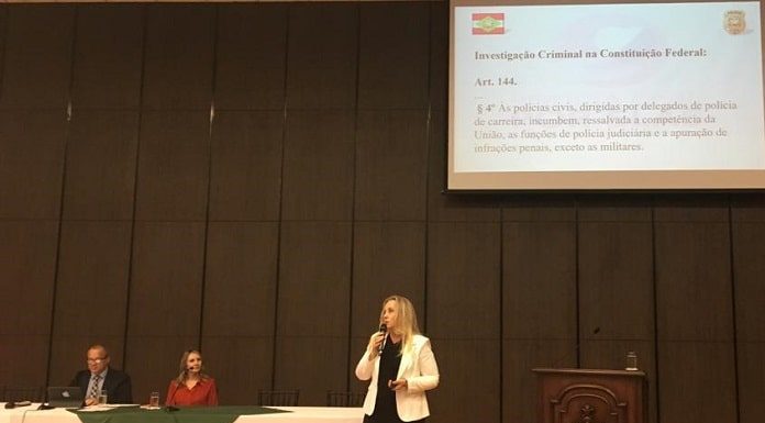 delegada em pé discursa com microfone na mão ao lado de pequena tribuna em palco com duas pessoas sentadas atrás de bancada