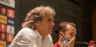 jorge jesus fala ao microfone gesticulando; há uma garrafa de água na bancada e uma pessoa atrás