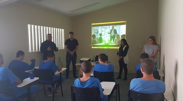 adolescentes sentados em carteiras e de costas para a câmera assistem adultos em pé falando com um projeção de slide na parede