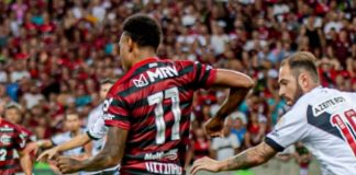 jogadores de flamengo e vasco se movimentando em campo com torcida ao fundo