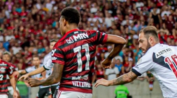 jogadores de flamengo e vasco se movimentando em campo com torcida ao fundo