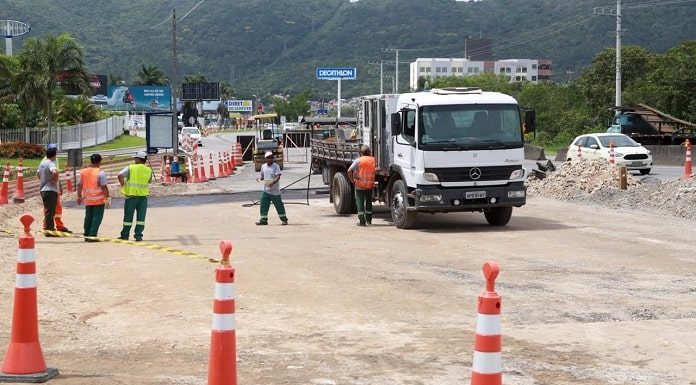 caminhões e operários sobre a pista onde há cones e montes de areia