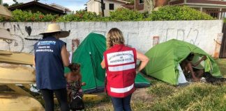 duas mulheres com uniformes da prefeitura vistas de costas conversando com moradora de rua ao lado de duas barracas em área de grama alta ao lado de um muro