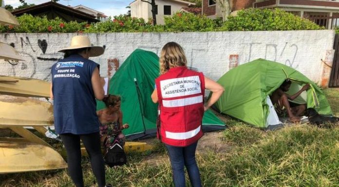 duas mulheres com uniformes da prefeitura vistas de costas conversando com moradora de rua ao lado de duas barracas em área de grama alta ao lado de um muro