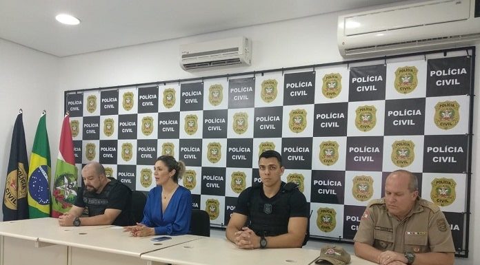 bancada com quatro pessoas: a delegada, um delegado, um policial municipal e um policial militar, com painel de logos da polícia civil e bandeiras ao fundo