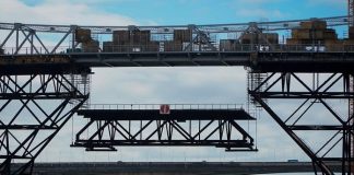 parte da treliça inferior à ponte sendo baixada no meio da estrutura com o mar abaixo e as outras pontes ao fundo