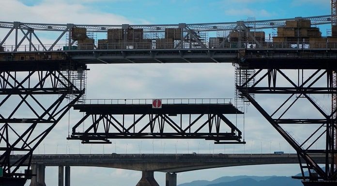 parte da treliça inferior à ponte sendo baixada no meio da estrutura com o mar abaixo e as outras pontes ao fundo