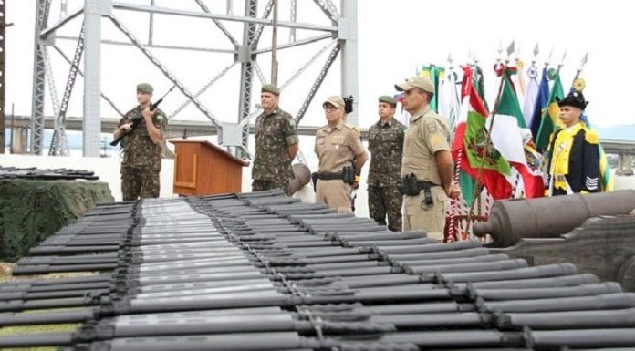 fuzis enfileirados em primeiro plano com militares do exército e da polícia ao fundo, bandeiras e uma estrutura da ponte hercílio luz