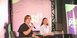 adeliana e marlene sentada em poltronas em um palco. adeliana segura um microfone; há um banner do partido ao fundo