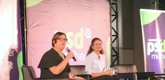 adeliana e marlene sentada em poltronas em um palco. adeliana segura um microfone; há um banner do partido ao fundo