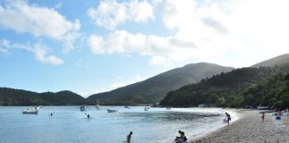 praia de mar calmo, cinco pessoas na praia, alguns barcos e morros de mata preservada ao redor