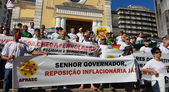 diversas pessoas com mesma camiseta na escadaria da catedral e faixas; à frente uma tem escrito "senhor governador, reposição inflacionária já"