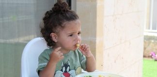 menina comendo em cadeirote infantil com prato de legumes a sua frente