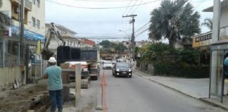 canteiro de obra em rua asfaltada com vala aberta, retroescavadeira e operário ao lado e carros passando na outra pista