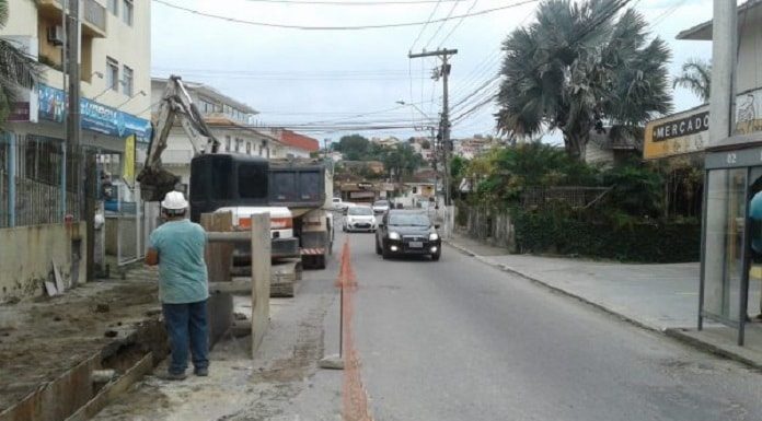 canteiro de obra em rua asfaltada com vala aberta, retroescavadeira e operário ao lado e carros passando na outra pista