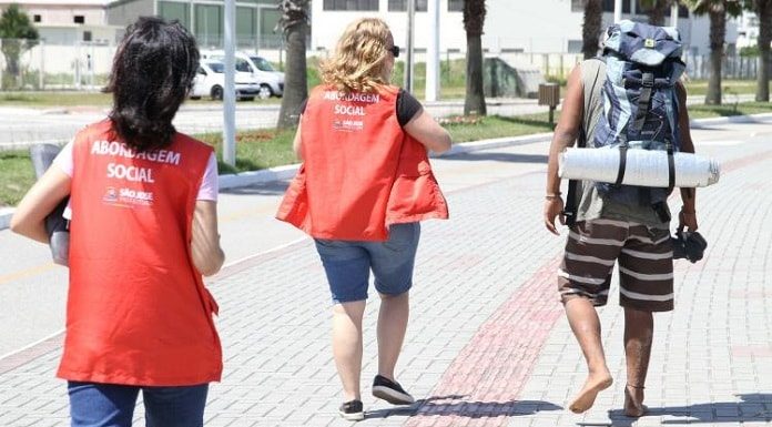 duas agentes do serviço social andando ao lado de homem com mochila e descalço na beira-mar de são josé