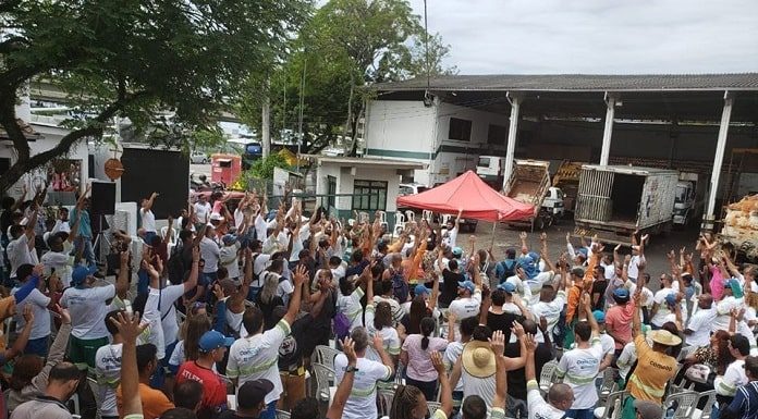 grande grupo de funcionários da comcap uniformizados com os braços para cima reunidos no pátio da companhia e gazebo do sindicato à frente com homem ao microfone