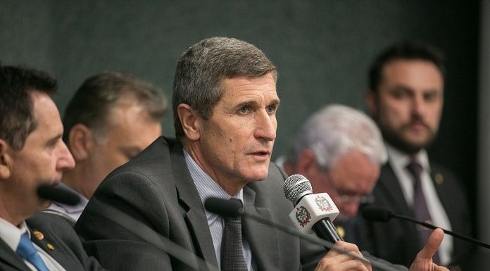 hassler falando ao microfone em uma bancada ao lado de outros homens de terno