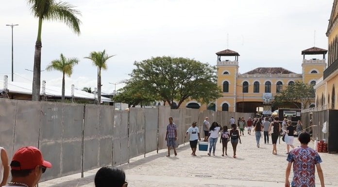 pessoas passando em rua de paralelepípedo entre o largo e a casa da alfândega, com tapumes de metal e mercado público ao fundo