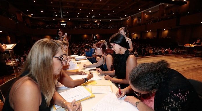 mesa onde mulheres estão dos dois lados, uma assinando papeis, outras entregando