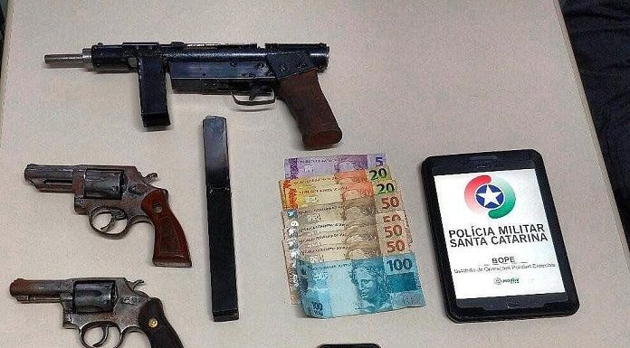 armas, celular e balas dispostas sobre uma mesa com tablet com logo da pmsc