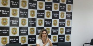 delegada sentada atrás de uma mesa falando e gesticulando e ao fundo painel com logos da polícia civil de sc