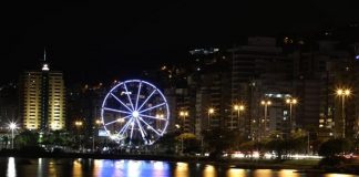 foto noturna da orla da beira mar com roda gigante em destaque e luzes do equipamento e da rua refletidas na água