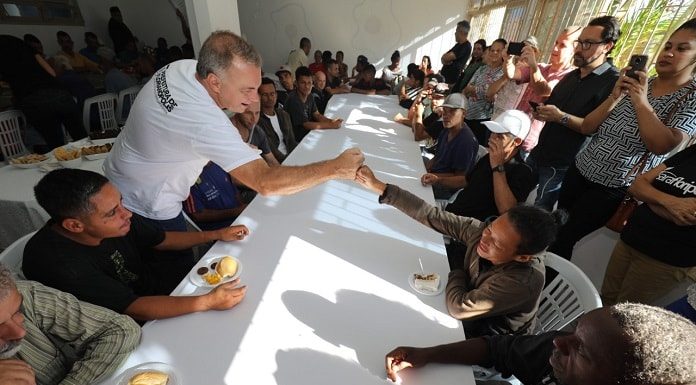 prefeito cumprimenta um homem do outro lado de uma grande mesa de refeitório cheia de gente sentada e em pé em volta