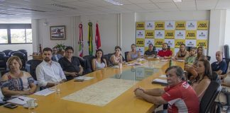 grupo de homens e mulheres reunidos em volta de grande mesa em sala que há painel da prefeitura ao fundo