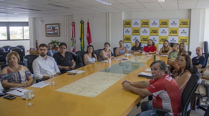 grupo de homens e mulheres reunidos em volta de grande mesa em sala que há painel da prefeitura ao fundo