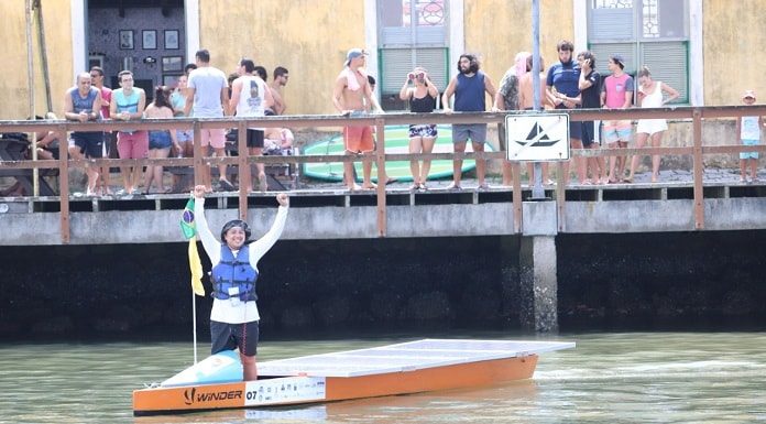 O piloto Marcos comemora o término da prova, de pé no barco em frente ao um pier com pessoas