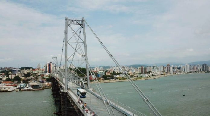 ônibus do transporte coletivo municipal de florianópolis passando pela ponte hercílio luz em foto aérea com o mar abaixo