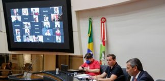 julio garcia na mesa do parlamento com dois homens ao lado e deputados em transmissão no telão