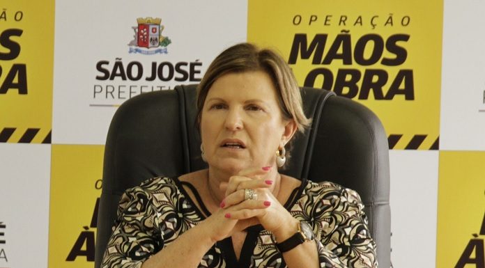 adeliana fala sentada na ponta de uma mesa com painel com logos da prefeitura ao fundo