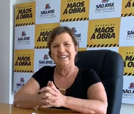 adeliana sentada atrás de mesa olha sorridente para câmera com painel da prefeitura ao fundo com logos