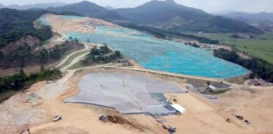 vista aérea do aterro sanitário de biguaçu, com boa parte de terra exposta, lonas por cima e máquinas circulando; montanhas ao fundo