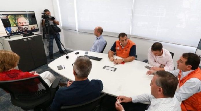 moisés e equipe reunidos ao redor de mesa olhando para uma tv onde está o ministro paulo guedes