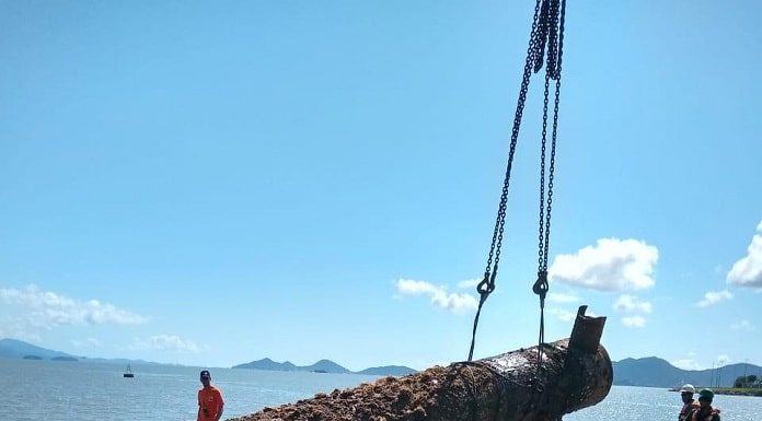 grande pilastra com mariscos sendo colada em balsa suspensa por cintas de um guindaste e operários em volta