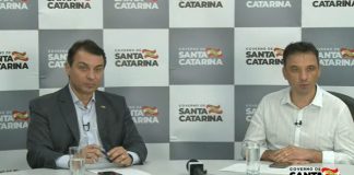 moisés e zeferino sentado atrás de mesa e no fundo painel do governo com logos