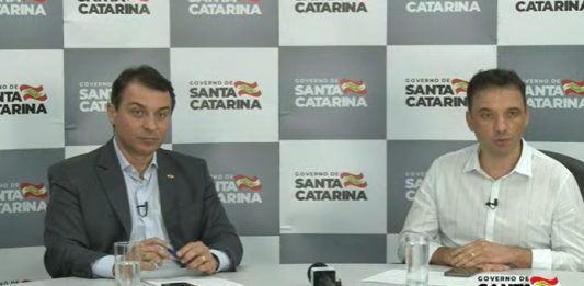 moisés e zeferino sentado atrás de mesa e no fundo painel do governo com logos