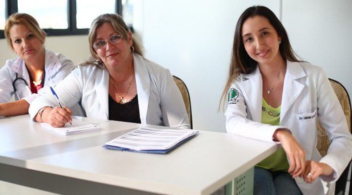 três médicas, duas de meia idade e outra mais jovem, sentadas atrás de mesa de consultório olham para a câmera