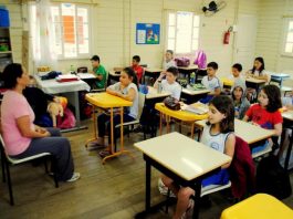 crianças sentadas em carteiras em sala de aula de madeira com professora à frente