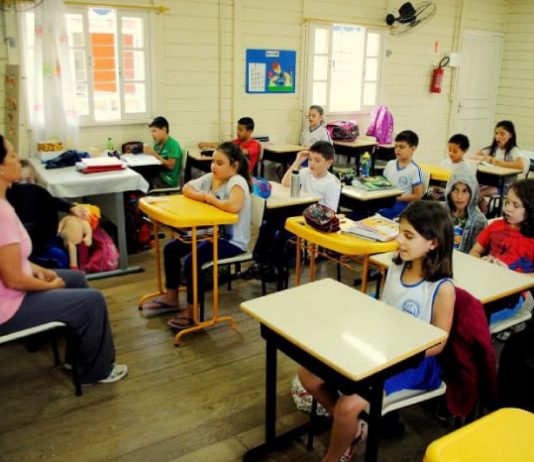 crianças sentadas em carteiras em sala de aula de madeira com professora à frente