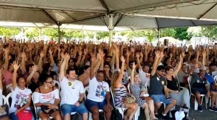 centenas de pessoas sentadas sob área coberta com os braços levantados