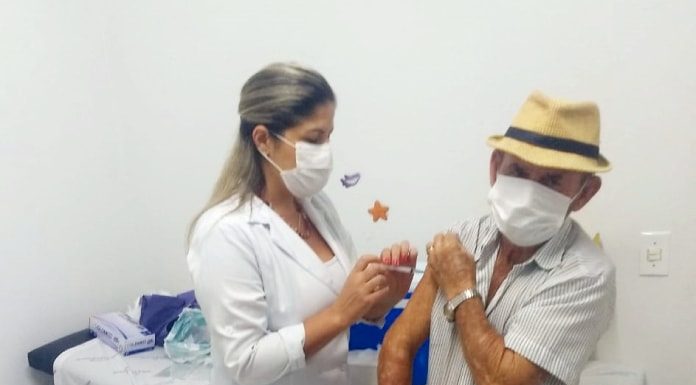 idoso com máscara no rosto usando chapéu de palha recebe vacina no braço esquerdo de uma profissional de saúde também usando máscara