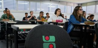 carteira vazia com logo da udesc bordado e alunos na sala de aula em volta