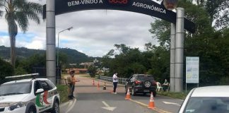 carro parado no sob o portal da cidade onde há cones na estrada e um homem de máscara ao lado do carro; carro da polícia; mais três pessoas em volta